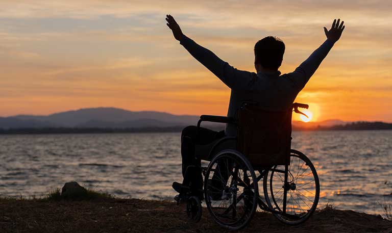 a man with raised arms in a wheelchair watching the sunset over a lake