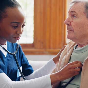 A woman in blue scrubs holds a stethoscope up to an elderly man's chest